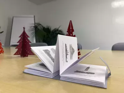 An open book displays architectural sketches on a light wood table, with burgundy decorative trees and a potted plant visible in the background during a holiday office display.