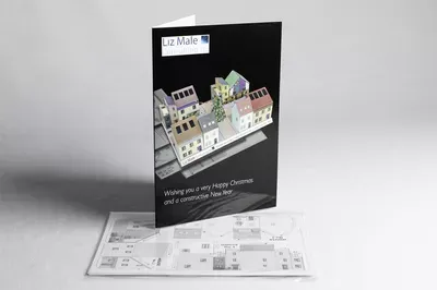 Black greeting card featuring a 3D architectural model of a street with buildings and storefronts, accompanied by unfolded card templates and construction diagrams on white paper below.