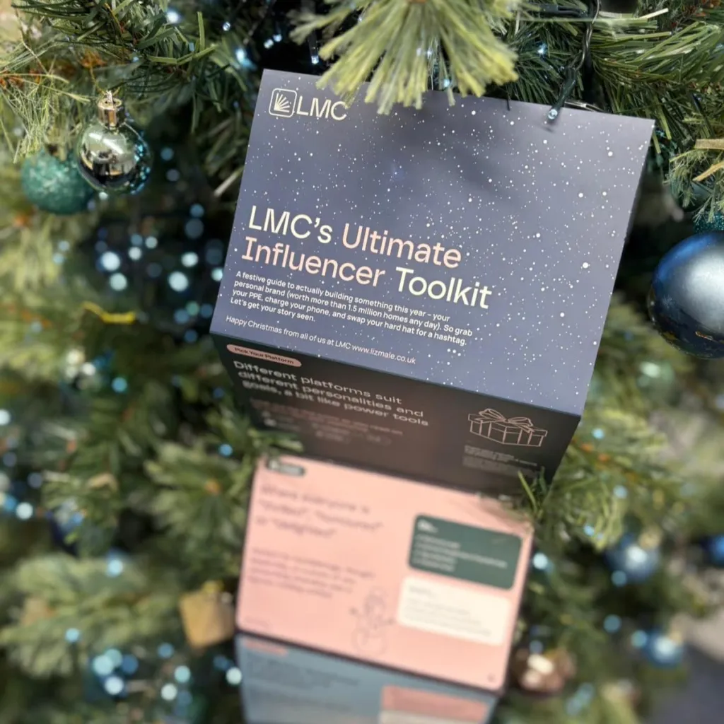 An open LMC's Ultimate Influencer Toolkit box lies on an evergreen branch surrounded by silver and blue Christmas ornaments, showcasing the product packaging with visible text and design elements.
