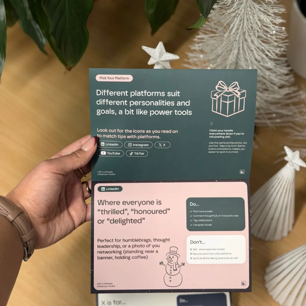 A person holds a printed marketing card about choosing social media platforms, featuring a teal section on different platforms and a pink section describing ideal audience engagement, with decorative holiday elements visible in the background.