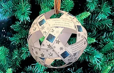 A white paper ball ornament decorated with small blue and teal snowflakes and text hangs from a gold ribbon in front of a Christmas tree with green branches.