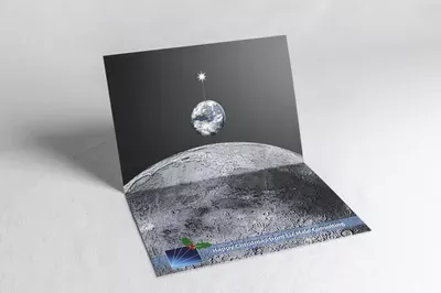 A pop-up Christmas card featuring a textured lunar landscape with a blue element, accompanied by a black panel depicting Earth and a star in space above the moon.