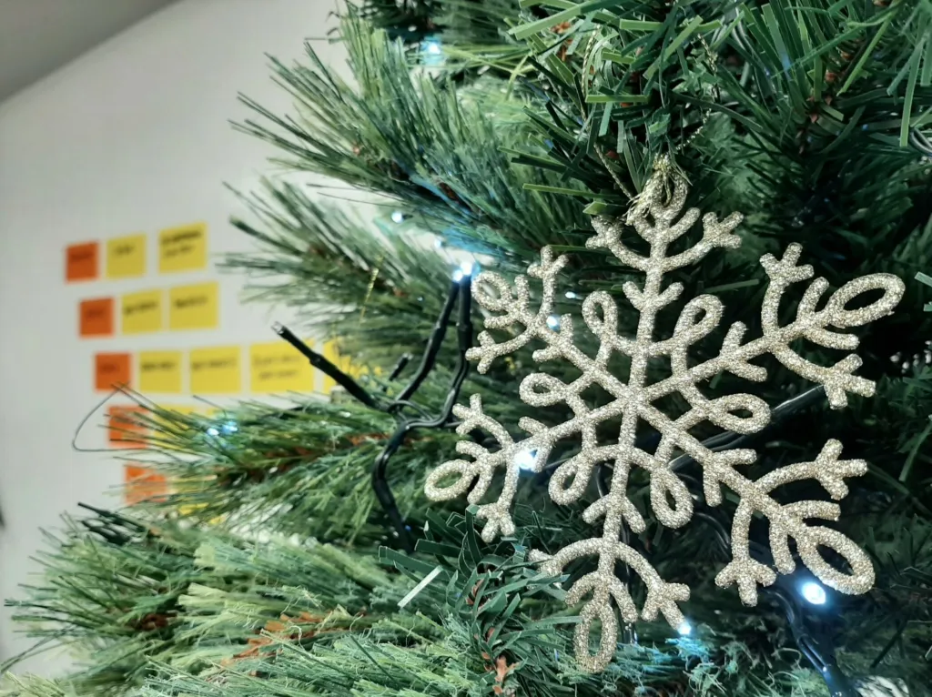 A glittery silver snowflake ornament hangs on a green tinsel Christmas tree branch. Blurred sticky notes in yellow and orange are visible in the background. The scene captures a festive holiday decoration setup.