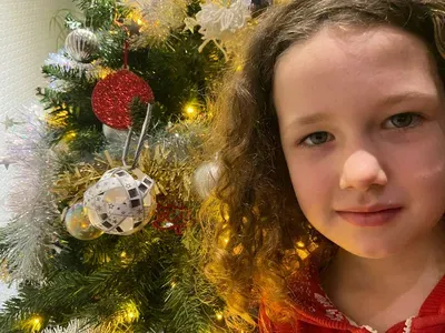 A young girl in a red shirt smiles at the camera next to a decorated Christmas tree adorned with gold lights, red ornaments, and a disco ball decoration.