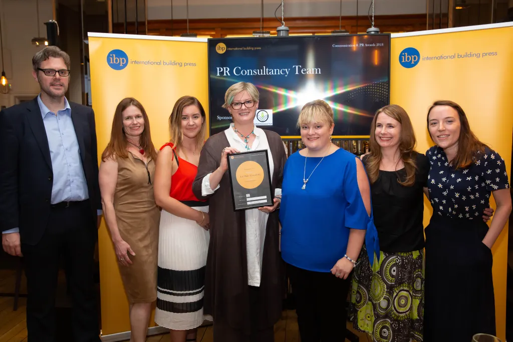 Seven professionals from the PR Consultancy Team stand together holding an award in front of branded yellow and dark banners at what appears to be a formal recognition event.