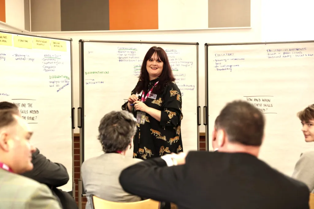A woman with dark hair wearing a black jacket with gold embroidery stands presenting to a group of seated attendees in front of whiteboards covered with notes and diagrams. She appears to be leading a workshop or training session in a professional setting.