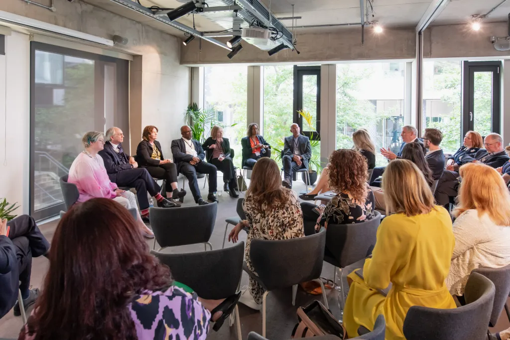 A diverse group of professionals seated in a circle during what appears to be a panel discussion or collaborative meeting in a modern office space with large windows and natural lighting.