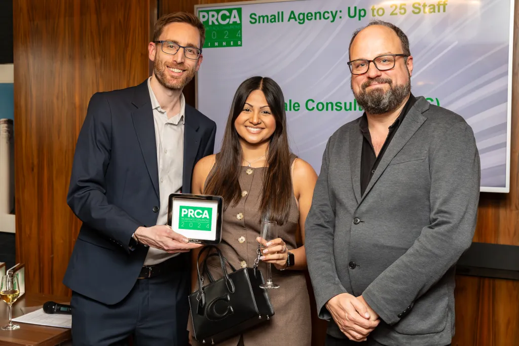 Three professionals stand together at an awards event, with the person on the left holding a PRCA award. A presentation screen behind them displays text about small agencies and PRCA branding.