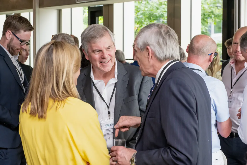 A woman in a yellow blouse shakes hands with an older man in a dark suit at a professional networking event, surrounded by other attendees wearing name badges indoors near large windows.