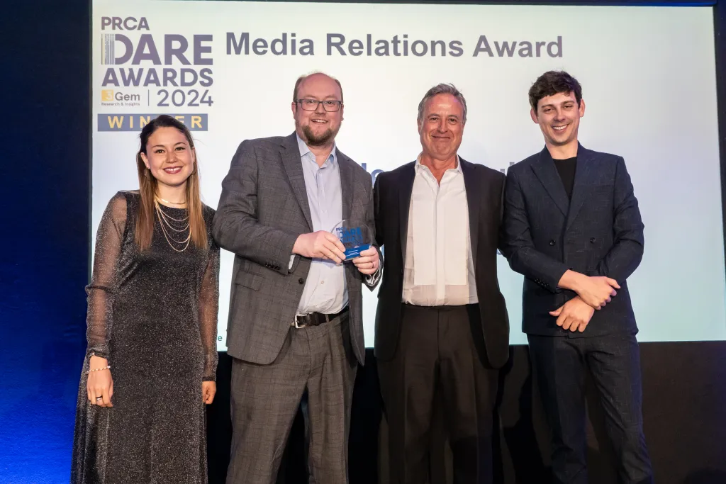 Four professionals pose together at the PRCA Dare Media Relations Award 2024 ceremony, with one man holding an award plaque. They stand in front of a backdrop displaying the event branding and sponsor logos.