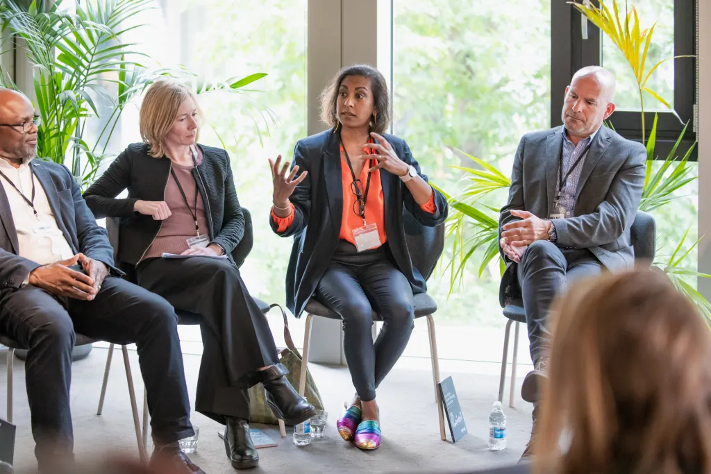 Four business professionals sit on chairs during a panel discussion in a bright, plant-filled indoor space. The woman in the center, wearing an orange lanyard, gestures while speaking to the group. An audience member is visible in the foreground listening to the panelists.