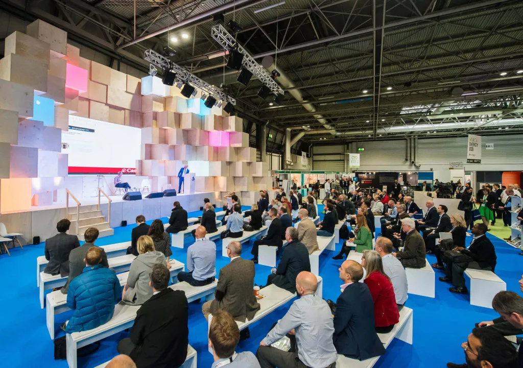 A large conference hall filled with attendees seated on blue flooring facing a colorful pixelated display wall and stage, with industrial ceiling and overhead lighting visible throughout the warehouse-style venue.