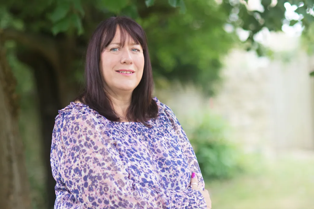 A woman with shoulder-length dark hair and bangs smiles while standing outdoors in front of trees. She wears a purple and cream patterned blouse. The background shows a blurred green garden setting.