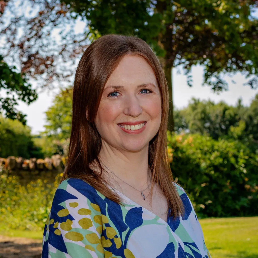 A woman with reddish-brown hair smiles at the camera while standing outdoors in front of green trees and grass. She wears a short-sleeved blue and green floral patterned top. The photo appears to be a professional headshot taken in natural daylight.