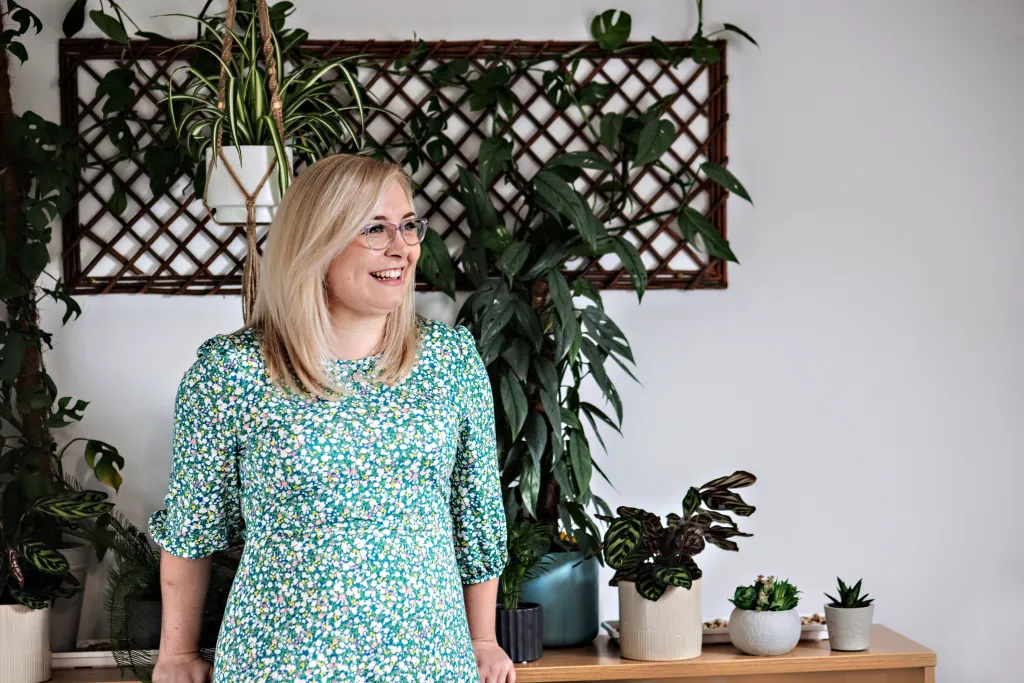A woman with blonde hair and glasses wearing a teal floral print shirt stands in an office environment surrounded by potted plants and a wooden lattice backdrop.