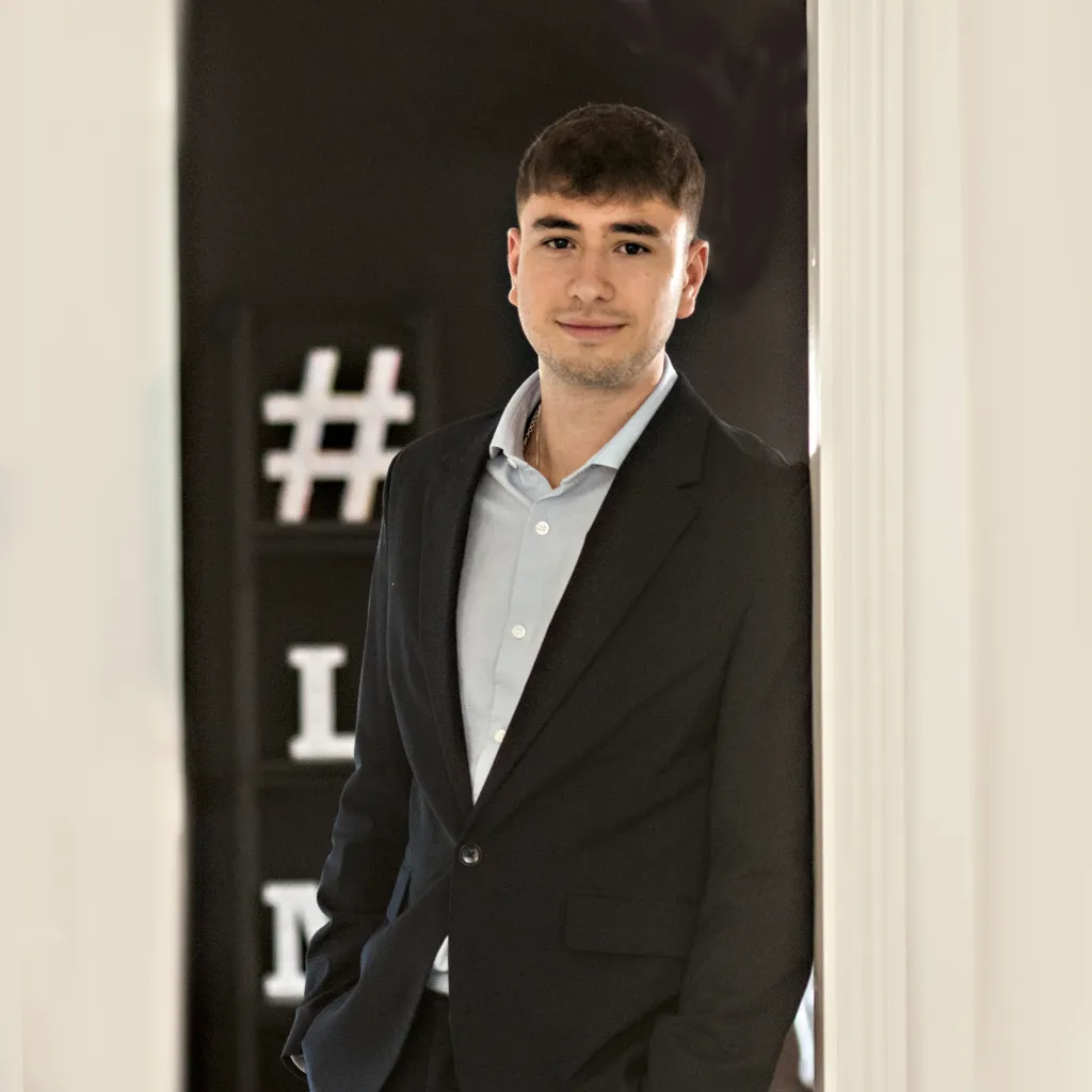 A young man with dark hair wearing a black blazer over a light blue shirt stands against a dark background with partial white text visible behind him.