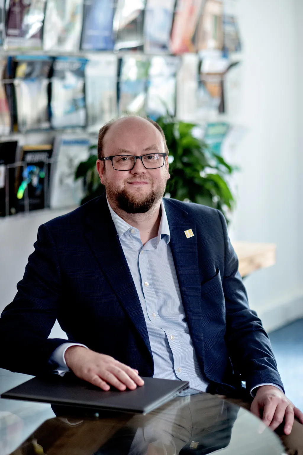 A man with glasses and a beard wearing a dark blazer and light blue shirt sits at a desk in a modern office, with a plant and displayed materials visible in the background.