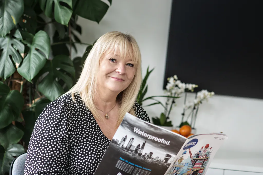 A woman with blonde hair and bangs wearing a dark polka dot blouse sits holding a magazine, smiling at the camera. She is surrounded by green plants and flowers in an office setting.