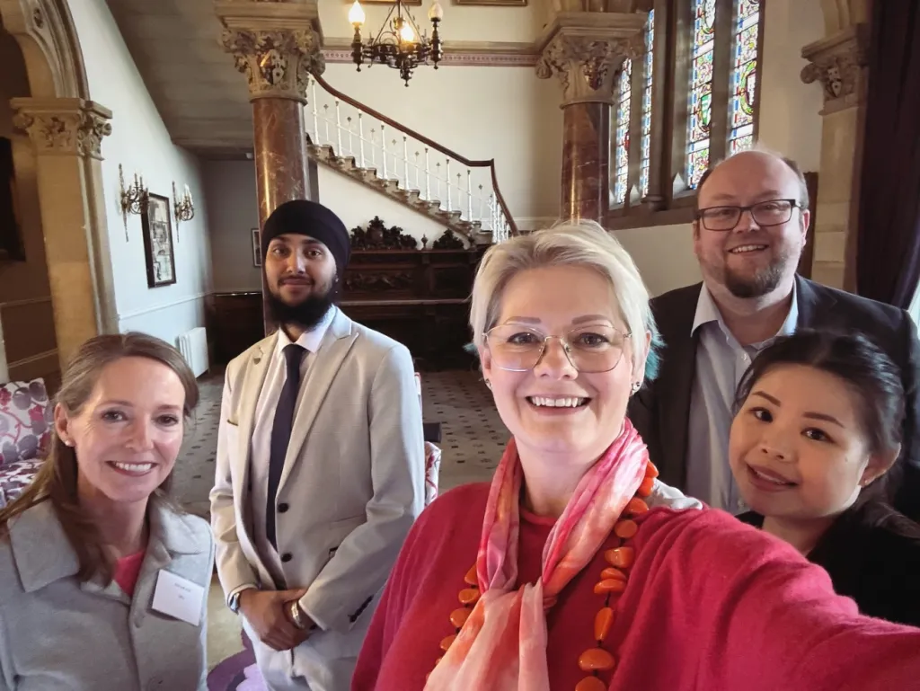Five team members from Liz Male Consulting pose together in an elegant hallway with classical architecture, ornate columns, a grand staircase, and a chandelier visible in the background.