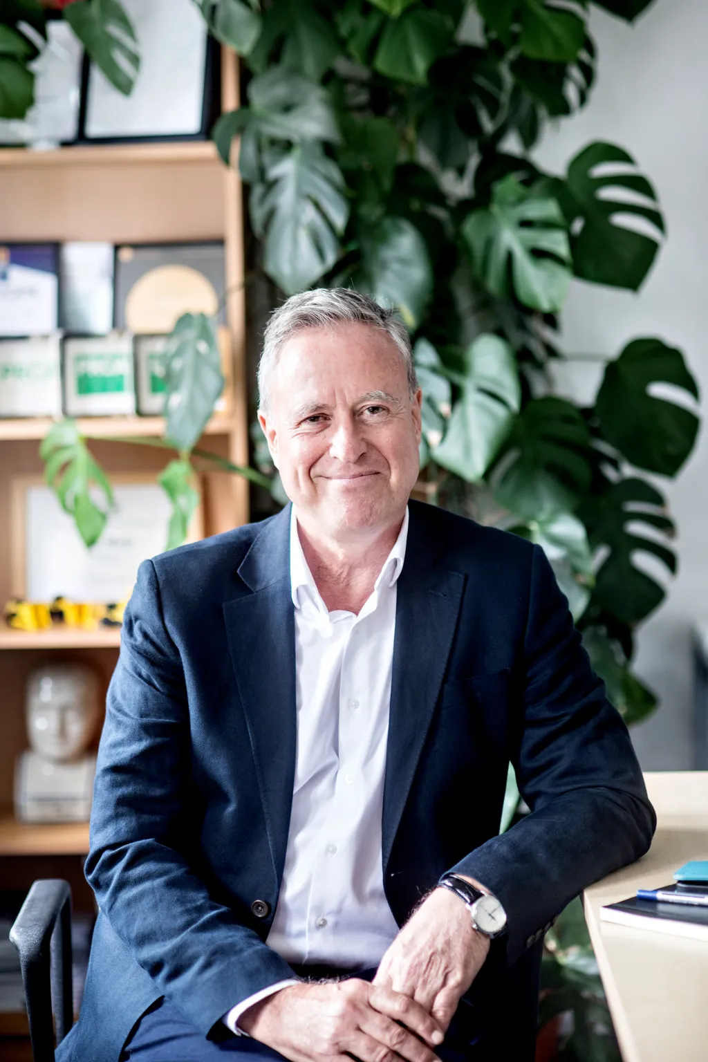 A professional man with gray hair wearing a dark blazer and white shirt sits in an office chair, smiling at the camera. Behind him is a wooden shelving unit decorated with potted green plants and office items. The setting suggests a modern, plant-filled workplace environment.