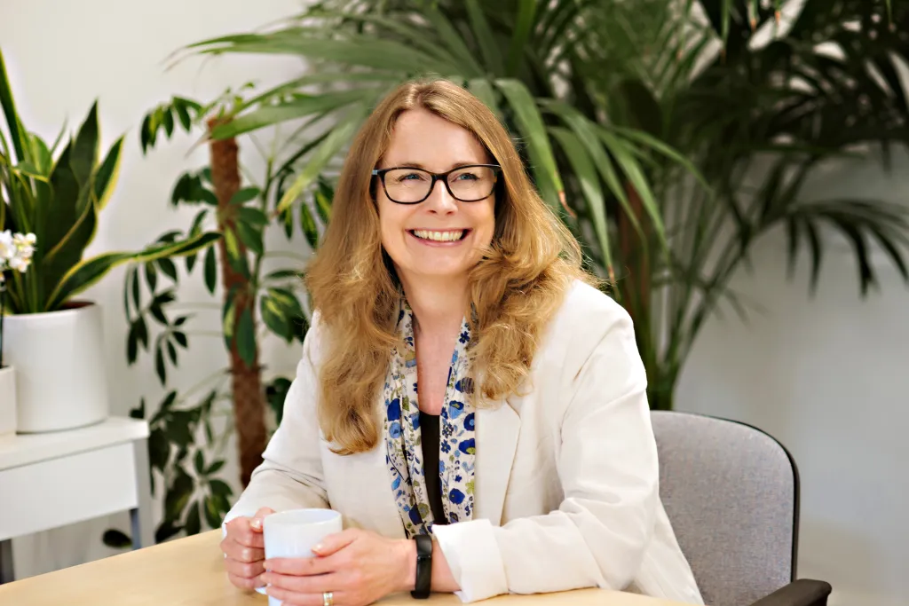A woman with shoulder-length strawberry blonde hair and glasses sits at a desk smiling at the camera. She wears a white blazer over a blue and white patterned blouse and holds a white mug. Green potted plants are visible in the background.