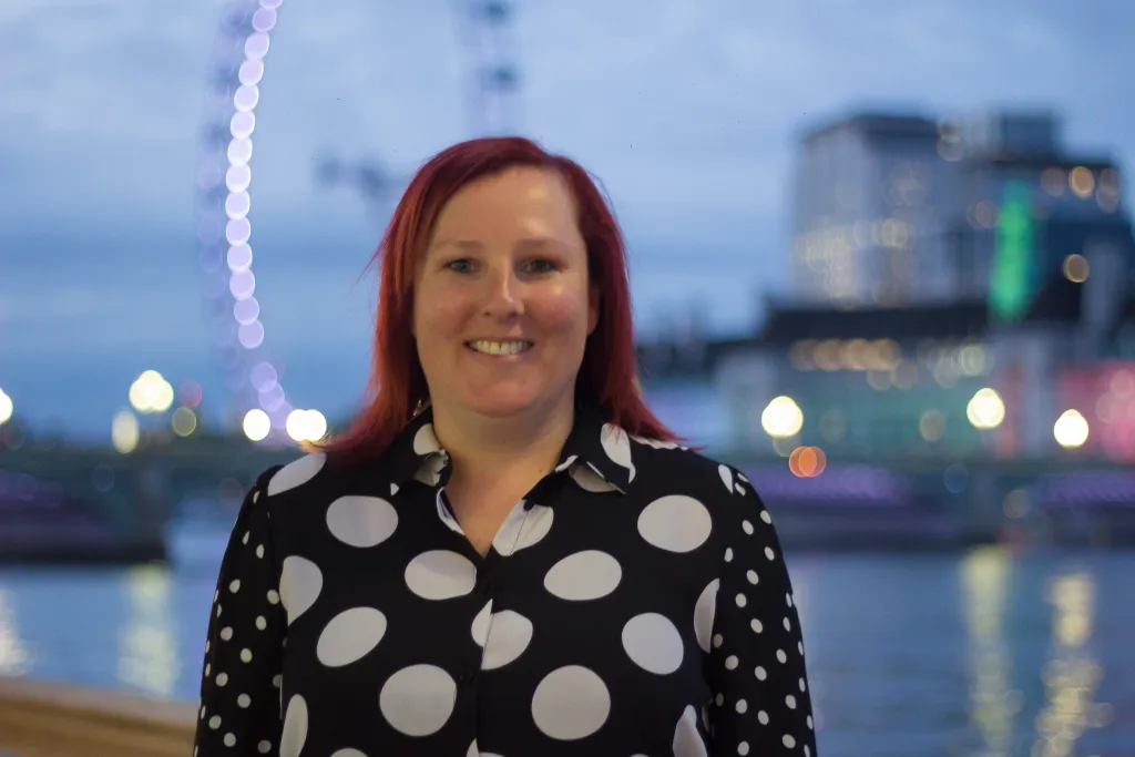 A woman with long reddish hair smiles at the camera while wearing a black and white polka dot blouse. Behind her is a twilight cityscape with illuminated buildings and water reflections.