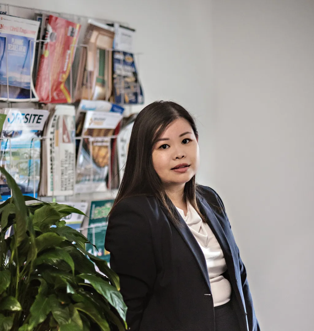 A woman with long dark hair wearing a dark blazer and white top stands in an office setting, looking upward thoughtfully. Behind her is a wall display with travel brochures and postcards, and a green plant is visible in the foreground.