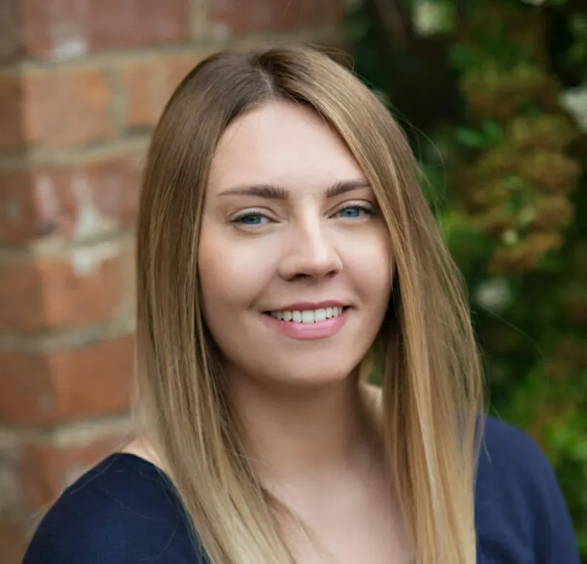 A smiling woman with shoulder-length light brown hair wearing a dark navy top, photographed against a brick wall with green foliage in the background.