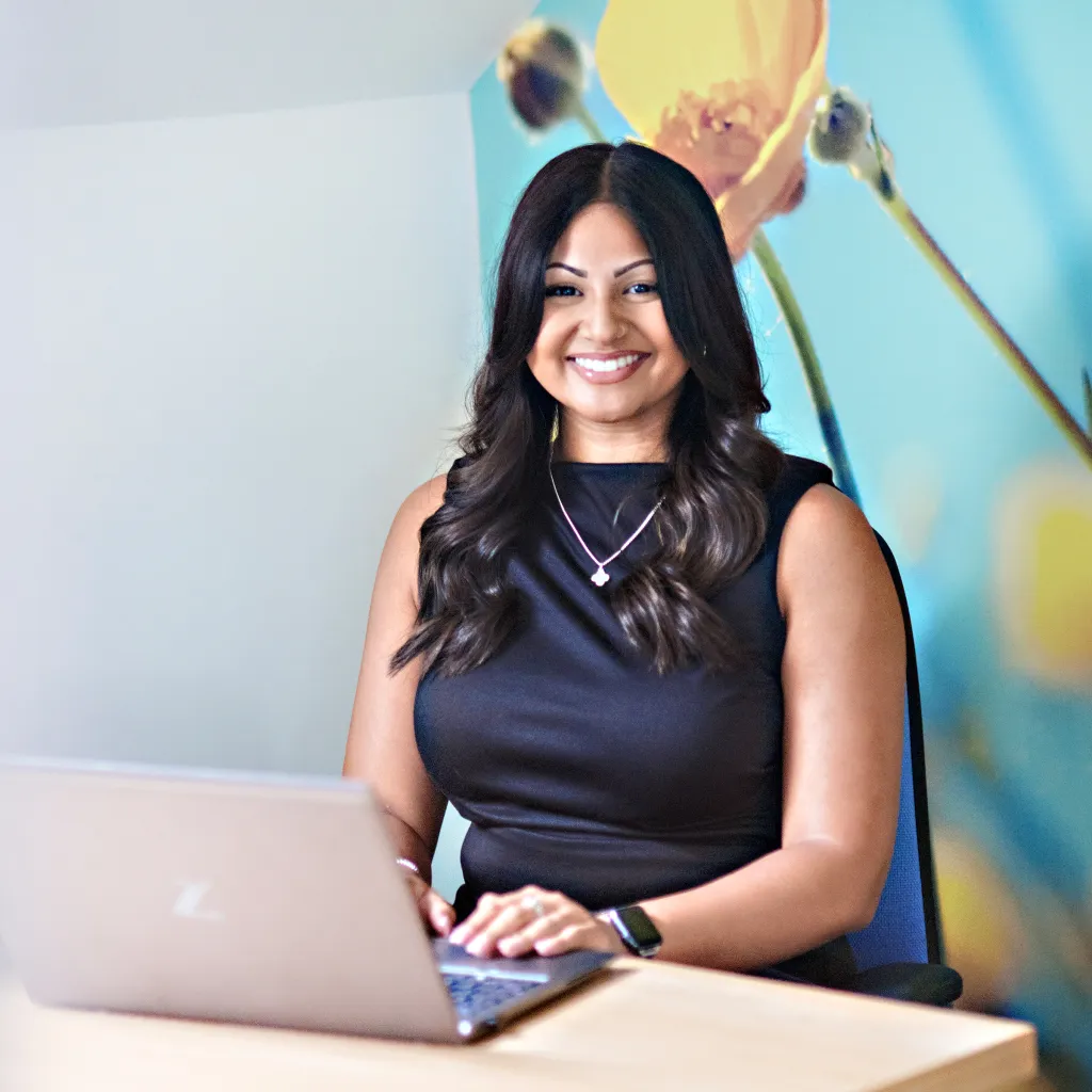 A woman with long dark hair wearing a black sleeveless top and necklace sits at a wooden desk with a laptop, smiling at the camera in a modern office setting with a blue wall and yellow floral artwork behind her.