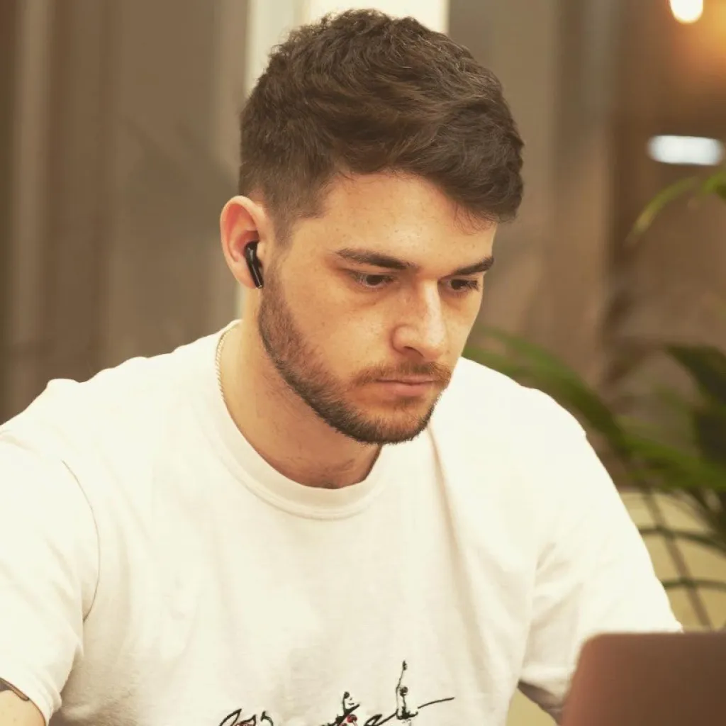 A young man with dark hair and facial hair wearing a white t-shirt and wireless earbud, focused on his laptop while working at a desk in a modern office setting.