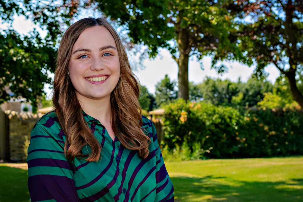 A smiling woman with long brown wavy hair wearing a green and navy striped long-sleeve shirt stands outdoors in front of trees and green grass.