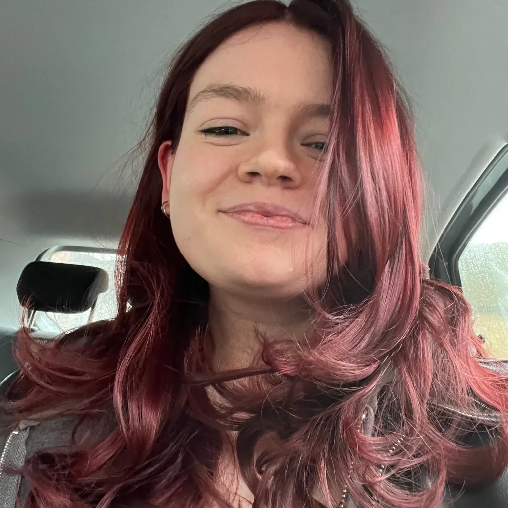 A woman with long, wavy burgundy hair and a subtle smile, photographed in a car during daytime. She is wearing dark clothing and looking toward the camera.