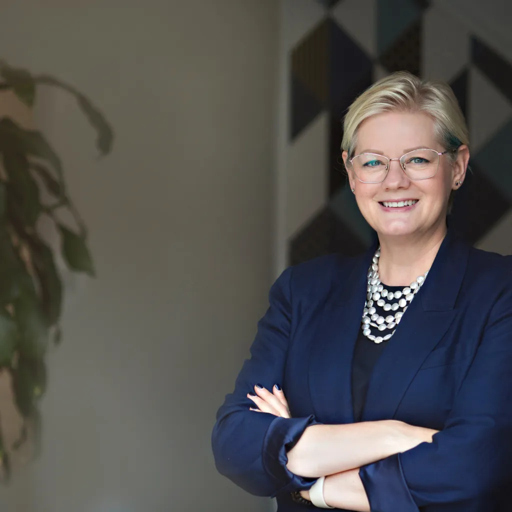 A professional woman with short blonde hair and glasses wearing a dark blue blazer and pearl necklace stands with arms crossed, smiling at the camera in an office setting with a plant visible in the background.
