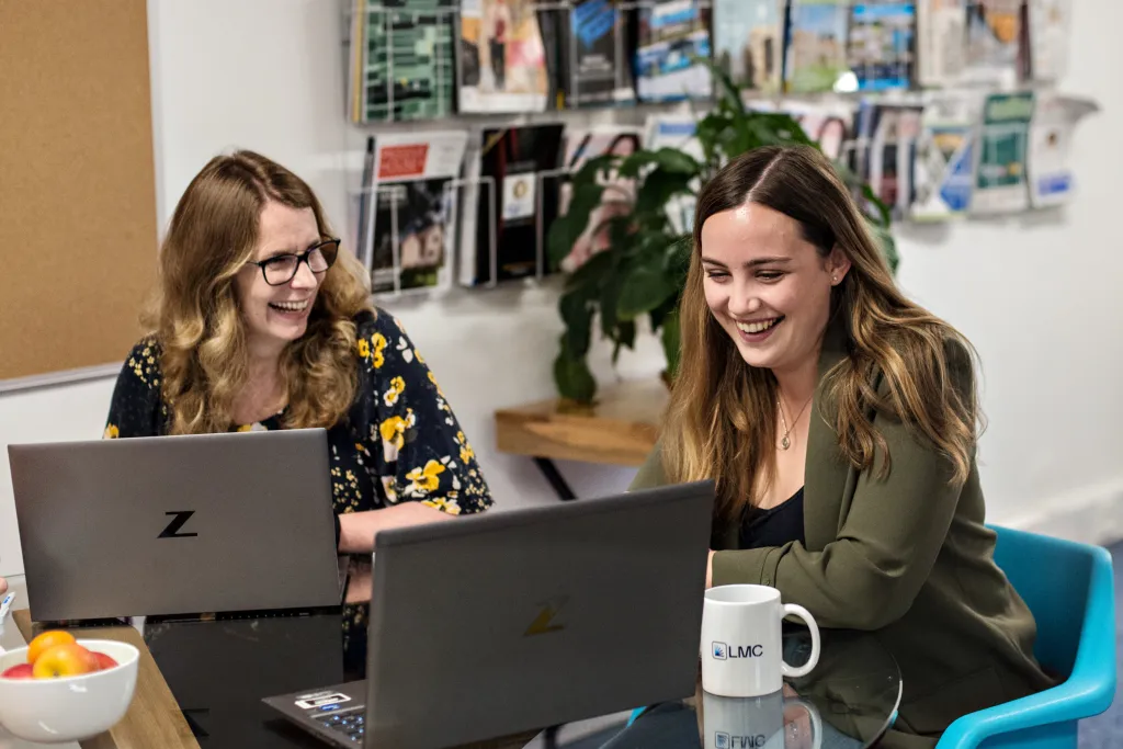 Two women sit at a desk working on laptops in an office setting. The woman on the left wears glasses and a dark floral top, while the woman on the right wears an olive green cardigan. They appear engaged and smiling while collaborating on work.