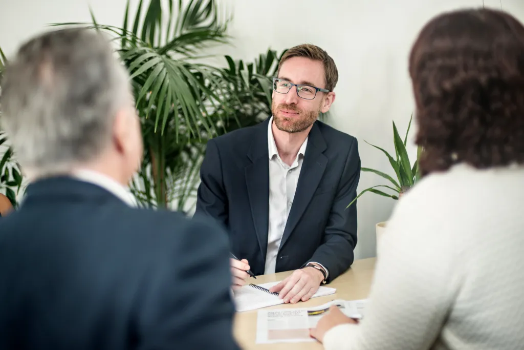Dan Gerrella, managing director of LMC, sits at a meeting room table, talking to another man and woman with their backs to the camera, with foliage in the background.