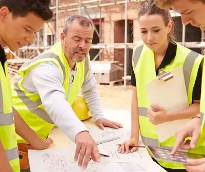 Construction workers in high-visibility vests review blueprints at a building site, with a brick structure under development visible in the background.