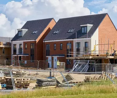 A residential construction site with three newly built brick homes featuring dark tile roofs, surrounded by scaffolding, building materials, and fencing during the framing and construction phase.