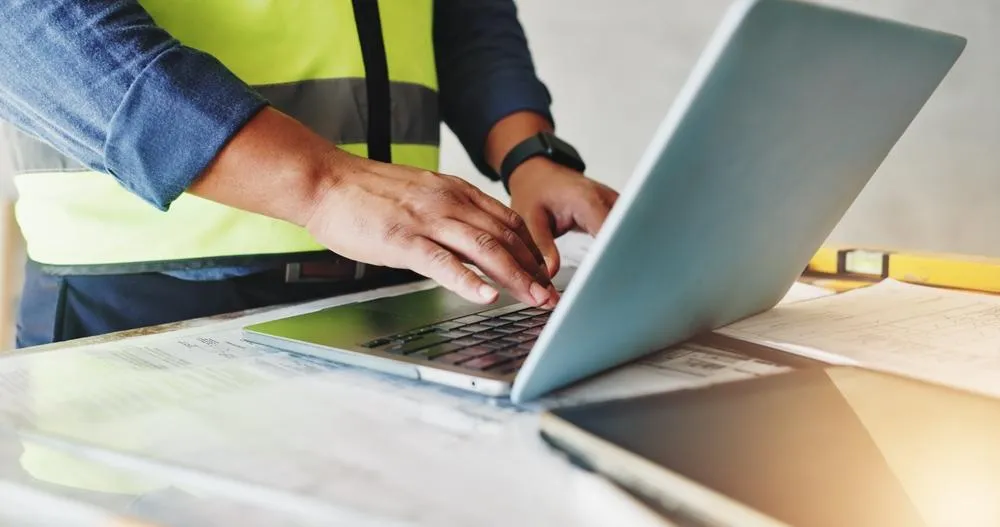 A construction worker wearing a high-visibility vest and hard hat types on a laptop while reviewing blueprints and documents on a wooden workbench.