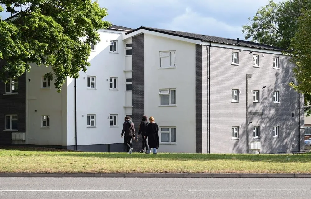 Modern four-storey residential apartment building with white and dark grey cladding, featuring regularly spaced windows and a contemporary design. Three people walk on the grass verge in front of the building alongside a paved road, with mature trees visible on either side.