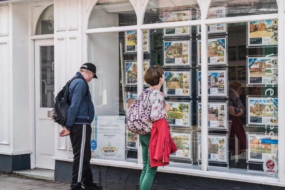 Two people stand outside a travel agency storefront, examining postcards and promotional materials displayed in the window. The person on the left wears a dark jacket and backpack with a hat, while the person on the right wears a patterned top and red cardigan with green tights. The storefront features white framing with multiple shelves of travel photographs and postcards.