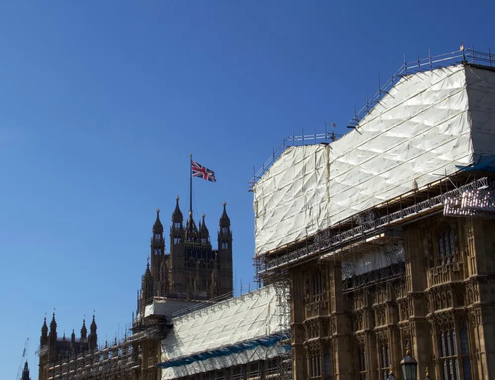Gothic towers of the Palace of Westminster with a Union Jack flag against a clear blue sky, with snow-covered scaffolding visible on adjacent historic stone buildings.