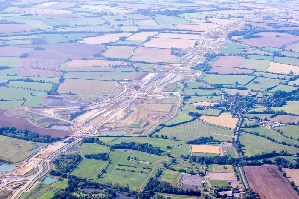 Aerial view of a patchwork of agricultural fields in various shades of green and brown, divided by hedgerows and tree lines, with a major highway interchange and infrastructure development visible cutting through the rural landscape.