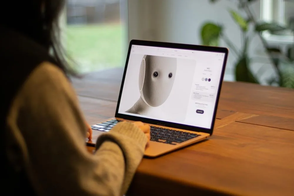 A person wearing a mustard-colored sweater sits at a wooden desk viewing a laptop screen displaying a 3D model of a minimalist white sculptural head or mask with simple dot features.