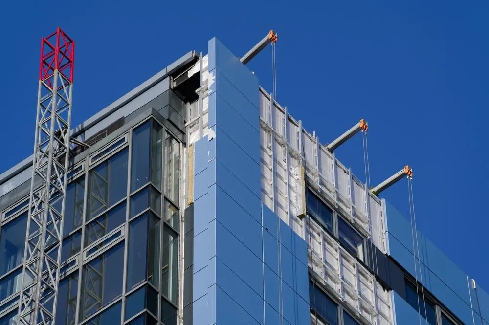 A modern multi-story building facade with large windows and white cladding panels against a clear blue sky, with construction cranes and safety netting visible on the exterior.