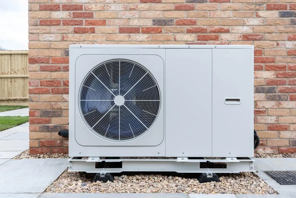 A white air source heat pump unit mounted on a concrete pad against a red brick wall, featuring a large circular fan vent on the left side and a control panel on the right.