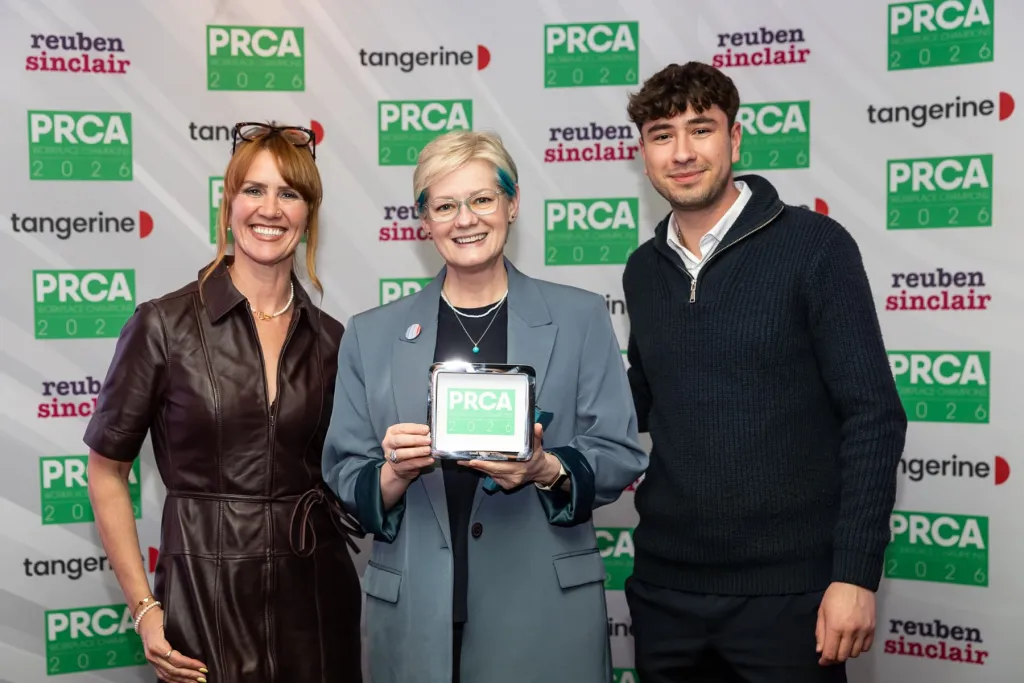 Three people stand in front a branded backdrop with PRCA, Reuben Sinclair, and Tangerine logos. The woman in the center holds a white award plaque while flanked by a woman on the left wearing a brown leather jacket and a man on the right in a dark sweater.
