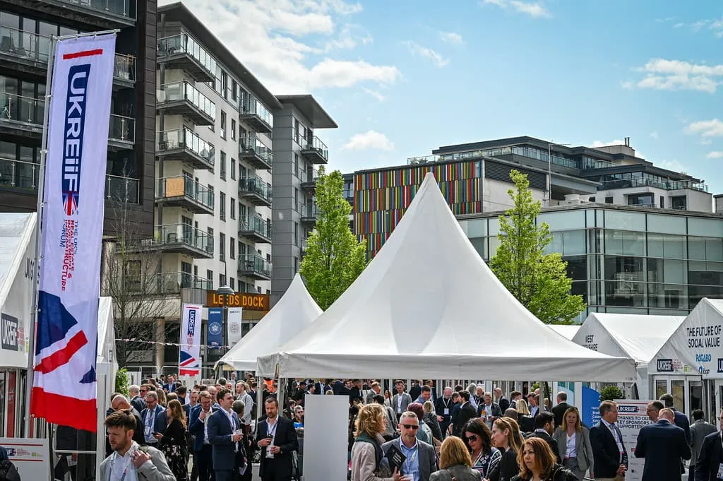 A large outdoor event with white tent structures in an urban setting, surrounded by multi-story buildings. Numerous attendees in business casual attire gather in the plaza area, with event signage and banners visible around the venue.