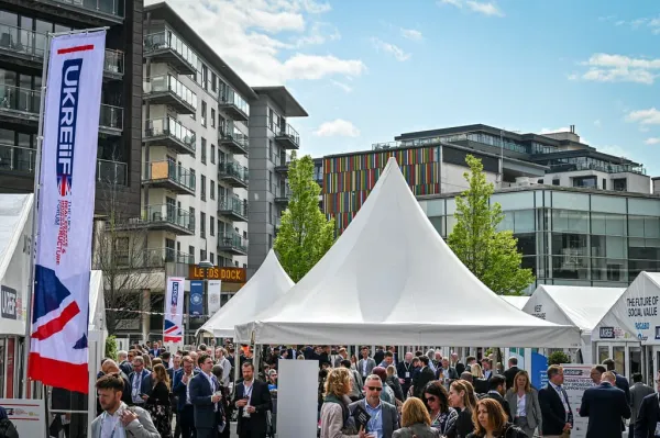 A large outdoor event with white tent structures in an urban setting, surrounded by multi-story buildings. Numerous attendees in business casual attire gather in the plaza area, with event signage and banners visible around the venue.