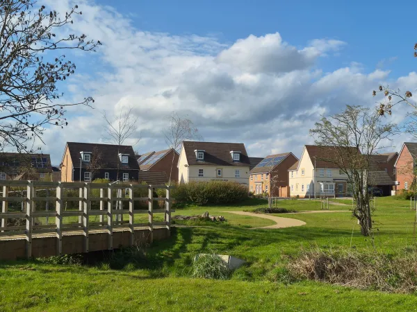A rural English village with brick and stone houses arranged around a green meadow, crossed by wooden fences and a small stream on an overcast spring day.