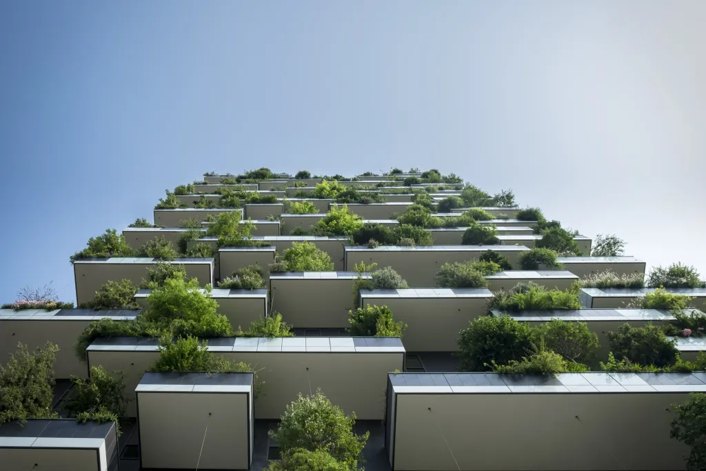 A modern residential building with multiple staggered levels, each featuring integrated green spaces with trees and vegetation growing from planted areas, demonstrating sustainable architecture and urban greening integration.